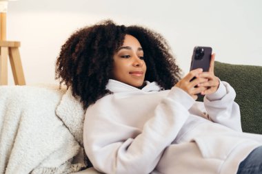 Afro saçlı genç bir kadın rahat bir koltukta dinleniyor, akıllı telefonunda rahatça kayıyor ve evinde huzurlu bir anın tadını çıkarıyor.
