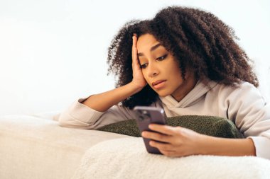 Stresli kadın evdeki koltukta uzanmış endişeli ve yorgun hissederken cep telefonu kullanıyor, kötü haberler okuyor ya da siber zorbalık yaşıyor.