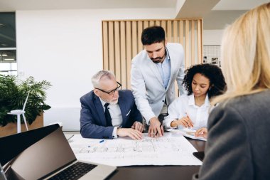 Mimarlar ve mühendisler proje projesinin planlarını inceliyor, minyatür rüzgar türbini modeliyle birlikte ofis ortamının planlarını ve özelliklerini tartışıyorlar. Buluşma konsepti