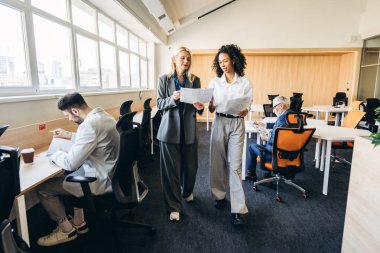 Modern bir ofiste yürüyen ve belgeleri tartışan iki iş kadını, iş arkadaşları özenle arka planda çalışırken işbirliği içinde çalışarak dinamik ve profesyonel bir ortam yaratıyorlar.