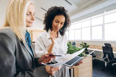 İki iş kadını bir proje üzerinde işbirliği yapıyor, finansal tabloları ve verileri dikkatle inceliyor ve modern ofis ortamında basılmış belgeler, takım çalışması ve uzmanlığı gösteriyor