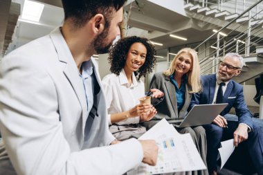 Çok ırklı iş adamları gayri resmi bir toplantıya katılıyor, pazarlama verilerini gözden geçiriyor ve modern bir ofis lobisinde raporları tartışıyor, işbirliği ve yenilikçi fikirleri teşvik ediyor. İş konsepti