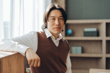 Beyaz gömlek ve kahverengi yelek giyen Asyalı genç bir adamın portresi, modern bir ofiste bir mobilyaya yaslanıyor.