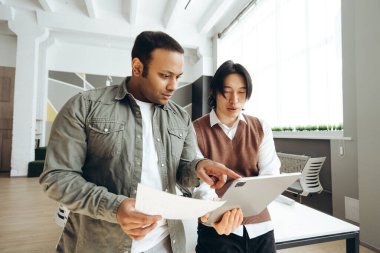 Dijital tablet ve belgeler üzerinde veri analiz eden iki genç pazarlamacı, modern bir ofiste birlikte çalışıyorlar.