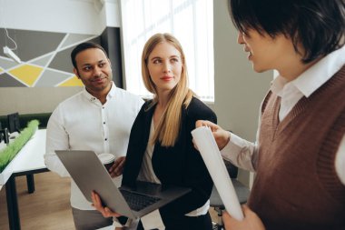 İş arkadaşları fikirleri paylaşıyor ve modern bir ofiste dizüstü bilgisayar ve belgeler kullanarak bir proje üzerinde birlikte çalışıyorlar.