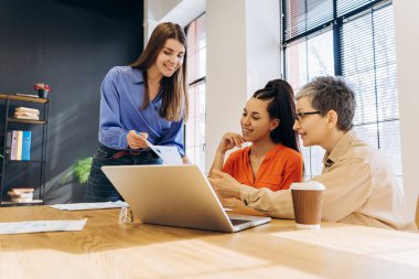 Güler yüzlü iş kadınları bir proje üzerinde işbirliği yapıyor, dizüstü bilgisayardaki verileri analiz ediyor ve modern ofis ortamındaki basılı belgeleri inceliyor, takım çalışması ve uzmanlığı teşvik ediyor