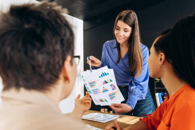 Renkli tablolar ve istatistiklere işaret eden genç iş kadını, modern ofiste yapılan verimli bir toplantıda şirket stratejisi ve performansını çok ırklı meslektaşlarıyla tartışıyor. İş konsepti