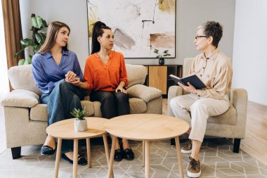Profesyonel bir psikologla terapi seansında el ele tutuşan ve sorunlarını paylaşan iki genç kadın bir kitap tutuyor ve modern ve parlak bir ofiste dikkatlice dinliyor.