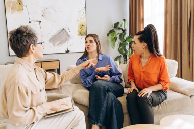 Bir terapi seansına rehberlik eden psikolog, anne ve kızı arasında sıcak bir oturma odası ortamında yaşanan aile çatışmasına arabuluculuk ederken, anlayış ve iletişimi geliştiren notlara işaret ediyor.