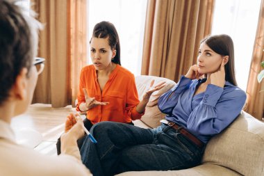 İki kadın çiftler terapisi sırasında bir koltukta otururken, biri kulaklarını kapatıp gözlerini kaçırırken, diğeri onunla tartışıyor ve psikolog da notlar alıyor.