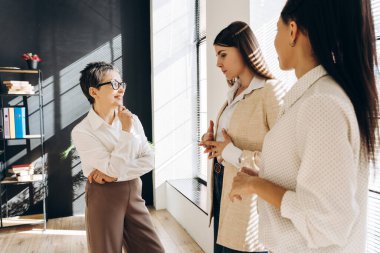 Üç iş kadını modern bir ofiste duruyorlar, bir tartışma içindeler, bir sonraki iş hamleleri için fikirlerini paylaşıyorlar ve stratejilerini paylaşıyorlar, camdan içeri akan doğal ışıkla banyo yapıyorlar.