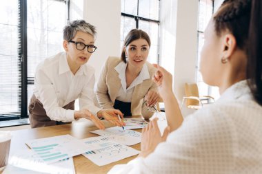 Üç iş kadını şirket performansını tartışıyor, finansal tabloları ve istatistikleri analiz ediyor, modern bir ofiste verimli bir toplantı sırasında bir proje üzerinde birlikte çalışıyorlar.
