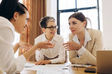 Üç iş kadını modern bir ofiste verimli bir toplantı sırasında grafik ve istatistikleri tartışıyor, takım çalışması, işbirliği ve veri odaklı karar verme mekanizmasını gösteriyor