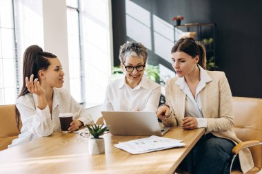 Üç iş kadını modern bir ofiste bir masada oturuyor, bir dizüstü bilgisayar kullanarak bir proje üzerinde işbirliği yapıyor ve iş hakkında konuşuyor, fikirleri paylaşıyor ve verileri analiz ediyorlar.