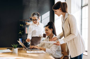 Bir proje üzerinde işbirliği yapan üç iş kadını, dizüstü bilgisayardaki verileri analiz ediyor ve parlak, modern ofis ortamındaki belgeleri inceliyor, takım çalışmasını ve üretkenliği teşvik ediyor