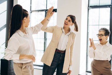 Üç iş kadını el çırpıyor, başarılı bir iş projesini veya modern ofis ortamında elde edilen başarıyı kutluyor.