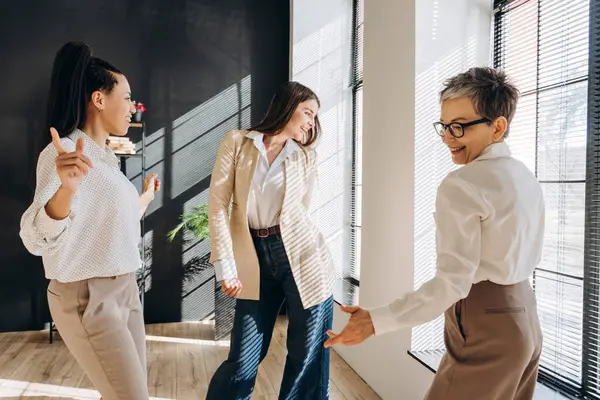 Üç mutlu iş kadını ofiste birlikte dans ediyor, tatillerinin tadını çıkarıyor ve başarılarını kutluyor, takım çalışmasını ve pozitif çalışma ortamını destekliyor.