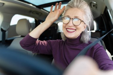Neşeli, gözlüklü, modern bir araba sürerken mutlu bir şekilde el sallayan güneşli bir günde özgürlüğü ve macerayı kucaklayan son sınıf öğrencisi bir kadın.