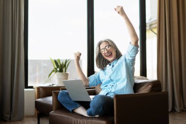 Evde dizüstü bilgisayar kullanırken heyecanla kollarını kaldıran mutlu yaşlı kadın, online galibiyeti, başarıyı veya iyi haberi kutluyor.
