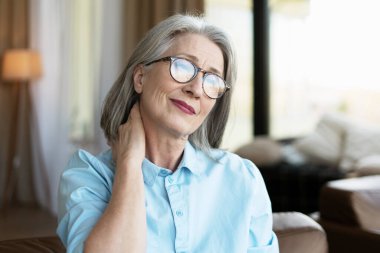 Gözlüklü olgun bir kadın boynuna dokunuyor. Uzun bir çalışma gününden sonra kasları kasılmış.