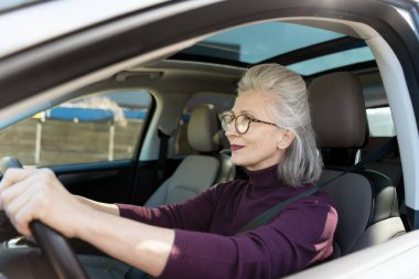 Mutlu olgun bir kadın arabasını sürüyor, şık gözlükler takıyor ve parlak bir şekilde gülümsüyor, açık yolların özgürlüğünün tadını çıkarıyor.
