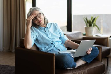 Olgun bir kadın migrenden muzdarip, alnına dokunmuş, kucağında bilgisayarla koltukta oturuyor.
