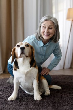 Gülümseyen son sınıf öğrencisi kadın oturma odasında diz çöküp köpeğine sarılıyor ve birlikte vakit geçiriyorlar.