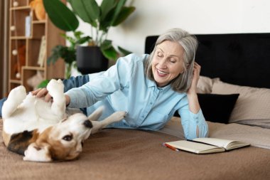 Gülümseyen olgun bir kadın evdeki yatakta rahat bir şekilde uzanırken neşeli av köpeğini kucaklıyor, neşe ve arkadaşlığın tadını çıkarıyor.