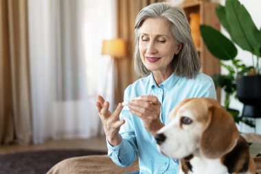 Yaşlı bir kadın kan şekerini evinde glukometreyle ölçüyor. Yanında da av köpeği oturuyor.