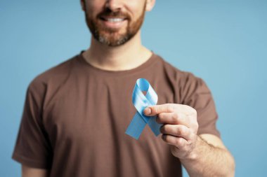 Gülümseyen adam gururla açık mavi bir kurdele sergiliyor, prostat kanseri farkındalığını sembolize ediyor ve bundan etkilenenlere sağlık ve destek veriyor.
