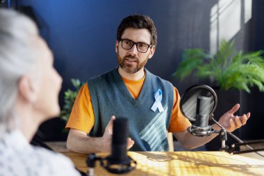 Açık mavi kurdele takan radyo sunucusu, kayıt stüdyosunda, prostat kanseri farkındalığından bahseden bir konukla röportaj yapıyor.