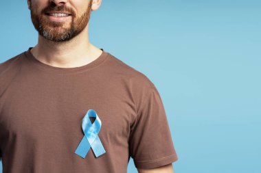Açık mavi kurdele takan, prostat kanseri farkındalığını temsil eden gülümseyen adam kahverengi tişörtünde gururla sergileniyor.