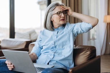 Üst düzey iş kadını elinde dizüstü bilgisayarla koltukta oturuyor migren ağrısı çekerken kafasına dokunuyor uzaktan çalışırken başı dönüyor ve yorgun hissediyor. Stres, sağlık konsepti