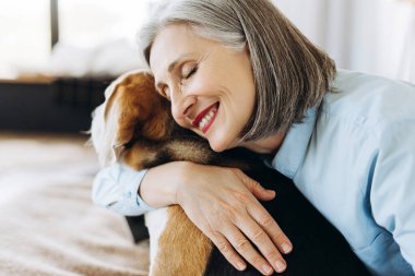 Gri saçlı, açık mavi tişörtlü, mutlu bir son sınıf öğrencisi, evinde köpeğini nazikçe kucaklıyor, sevgisini, ilgisini ve özel bir bağ anının tadını çıkarıyor.
