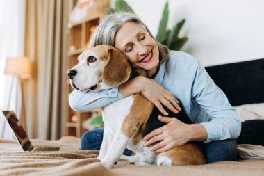 Mutlu, olgun, gözleri kapalı bir kadın, evinde dizüstü bilgisayarla yatağında oturmuş köpeğine sarılıyor. Birlikte vakit geçiriyorlar, sevgi ve özen gösteriyorlar.