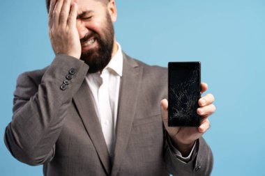 Umutsuz iş adamı, elinde bozuk bir akıllı telefon ekranını tutarken ağlıyor. Mavi arka planda hayal kırıklığını ve umutsuzluğu ifade ediyor.