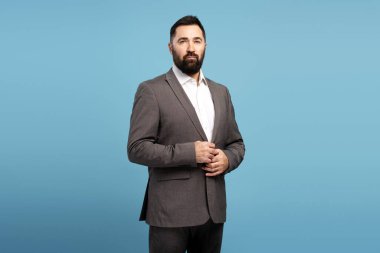 Kendine güvenen bir iş adamının portresi mavi arka planda dururken elbisesini düzeltiyor, profesyonelliği ve başarıyı aktarıyor.