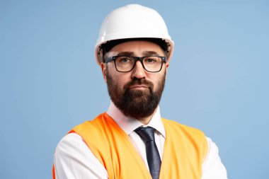 Koruyucu iş kıyafeti giyen kendine güvenen bir inşaat mühendisinin portresi mavi arka planda ciddi bir ifade sergiliyor.