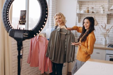 Elinde iki tişört olan ve video kaydeden gülümseyen iki bayan arkadaşın portresi. Evde odada dikiliyorum. Çevrimiçi kıyafet satışı sosyal medya