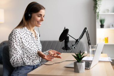 Gülümseyen kadın modern bir ev stüdyosunda mikrofon ve kulaklık kullanarak ilgi çekici bir ses yayını kaydediyor.