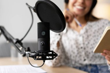 Podcaster, profesyonel mikrofon kullanarak bir podcast bölümünü kaydederken gülümsüyor ve bir not defterinden notlar okuyor.