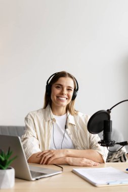 Podcaster kulaklık takıyor ve mikrofona konuşuyor, rahat bir stüdyo ortamında ses içeriğini kaydediyor.