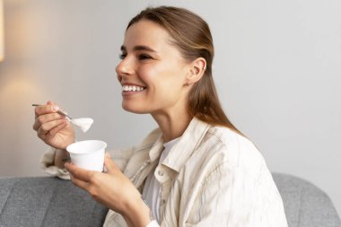 Kaşıkla plastik bardaktan yoğurt yiyen, kanepede oturan, sağlıklı kahvaltı ve atıştırmalıkların keyfini çıkaran mutlu bir kadın.