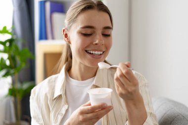 Plastik bardaktan yoğurt yiyen, oturma odasındaki rahat koltukta oturan mutlu genç kadın.