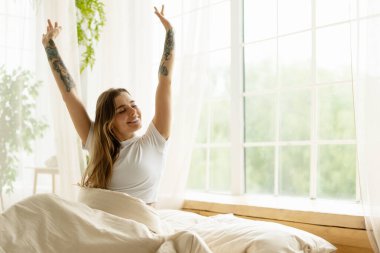 Mutlu kadın yatakta uzanıyor, sabah ışığını kucaklıyor ve enerji ve pozitiflikle dolu yeni bir günün tadını çıkarıyor.