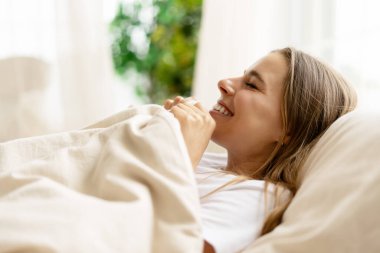 Mutlu bir kadın yatakta uyanıyor, neşeyle gülüyor, sıcak bir battaniye tutuyor, yatak odasındaki parlak sabah ışığının tadını çıkarıyor.