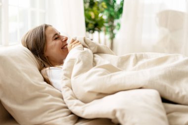 Sıcak battaniyelerin altında yatarken mutlu bir kadın gülüyor. Sıcak ve rahatlatıcı huzurlu bir sabahın tadını çıkarıyor.