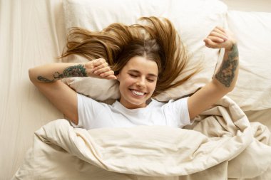 Dövmeli genç bir kadın uyanıyor ve yatakta geriniyor, mutluluk ve rahatlama yayıyor, yeni bir günün taze enerjisini kucaklıyor.