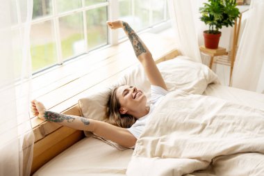 Sabah uyandıktan sonra yatakta uzanan mutlu genç bir kadın olasılıklarla dolu yeni bir günün mutluluğunu kucaklıyor.