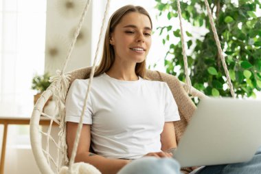 Evden çalışan, rahat salıncak koltuğunun keyfini çıkaran ve iş ve eğlence için dizüstü bilgisayar kullanan mutlu bir serbest yazar.